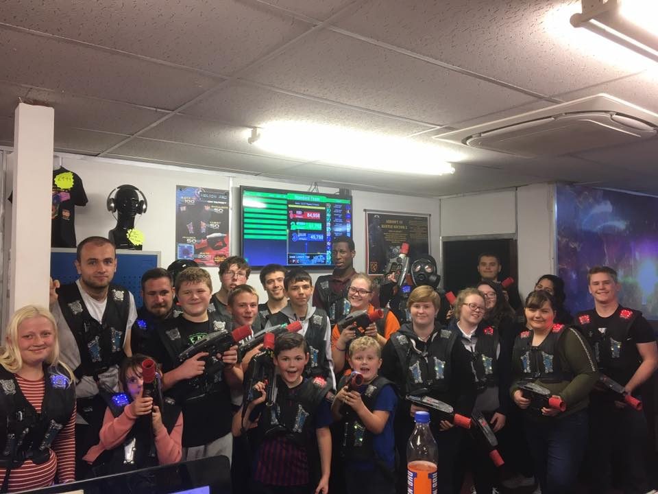 A big group of Puzzles members in laser tag vests in front of the scoreboard