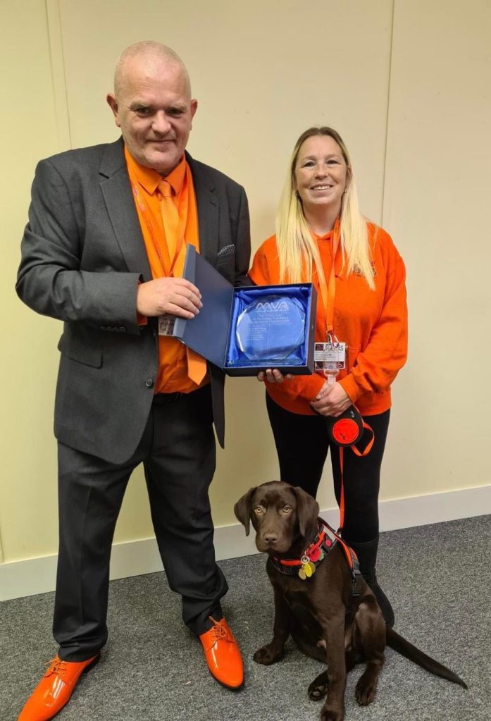 Paul Harper holding the MVA award with a colleague in a Puzzles orange hoodie and a chocolate Labrador