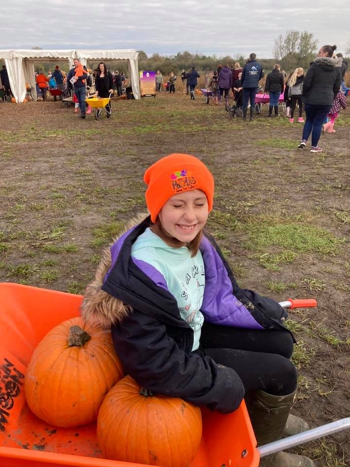 A smiling girl in an orange Puzzles beanie sitting in a wheelbarrow with pumpkins