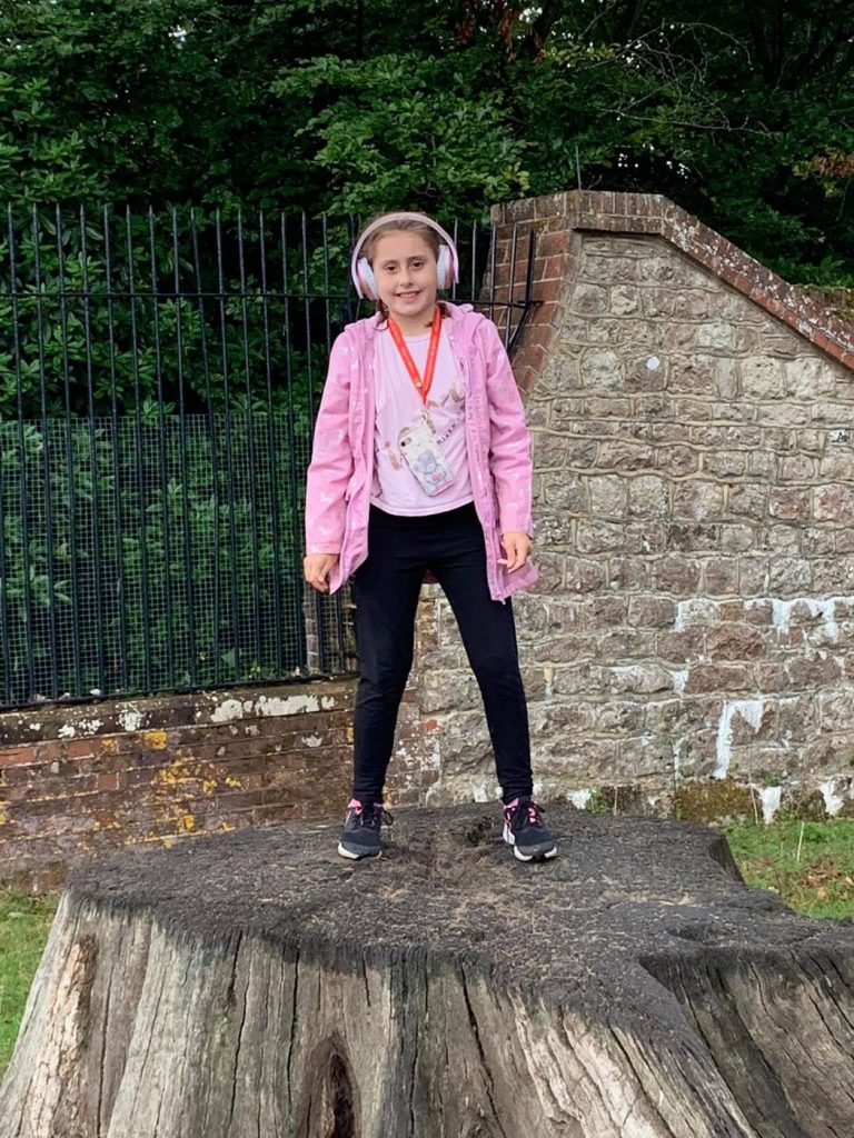 A girl wearing ear defenders standing on a large tree stump on a day out